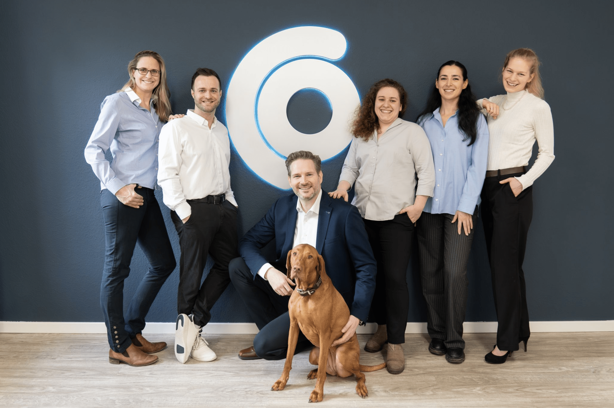 Oculus-Team mit Bürohund vor dem Firmenlogo – sechs Mitarbeitende lächeln in die Kamera